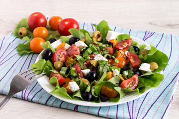 On a white plate is a fresh mouth-watering vegetable salad with tomatoes, olives, lettuce, feta and cashews.