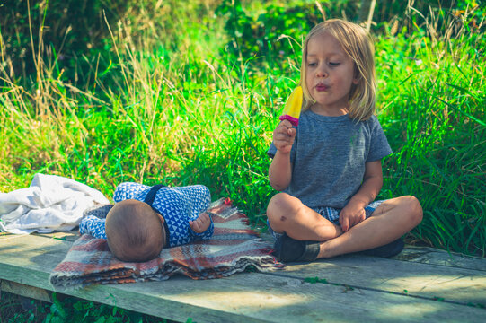 Preschooler eating ice lolly in garden wiith his baby sibling