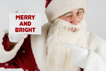Santa Claus holds a card with the text in his hands - MERRY AND BRIGHT