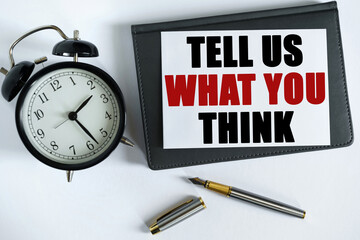 On the table there is a clock, a pen, a notebook and a card on which the text is written - TELL US WHAT YOU THINK