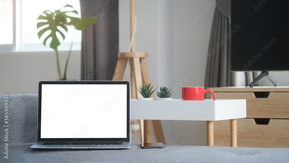 Poster an open laptop computer with blank screen and a smartphone are putting on sofa in living room.