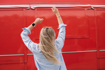 Rear view of woman standing against red wall with raised arms and blonde hair fluttering in the wind. Relaxing outdoors concept.