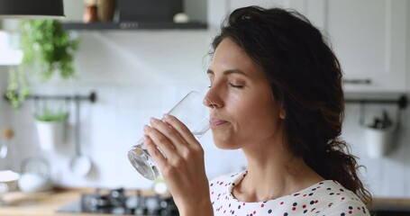 Thirsty smiling 35s woman standing alone in domestic kitchen start new day with healthy life habit, holding glass drinking clean mineral natural still water close up view. Lifestyle healthcare concept - Powered by Adobe
