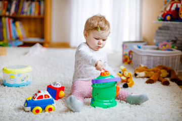 Happy joyful baby girl playing with different colorful toys at home. Adorable healthy toddler child having fun with playing alone. Active leisure indoors, nursery or playschool.