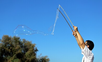 a boy who is making a big soap bubble with two sticks and a rope with a tree in the background and a beautiful blue sky