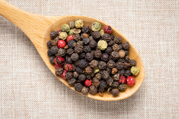 Pepper in a wooden spoon on light brown tablecloth. Pink and black peppercorn close-up. Colorful peas heap. Multicolor hot pepper seeds.