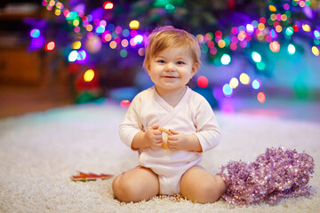 Fototapeta na wymiar Adorable baby girl holding colorful lights garland in cute hands. Little child in festive clothes decorating Christmas tree with family. First celebration of traditional holiday called Weihnachten