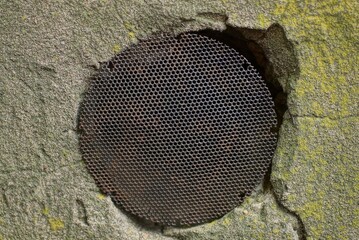black round iron grate in rust on the hole in the hood in the gray concrete wall outside