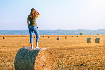 Naklejka na ściany i meble Back of a tall woman standing on a hay bale in harvested field and looking in the distance towards the horizon.