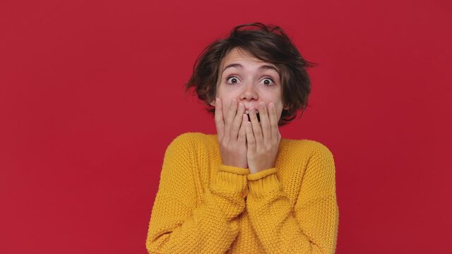 Happy fun beautiful young woman 20s in yellow sweater posing isolated on red wall background in studio. People emotions lifestyle concept. Looking camera shocked surprised say wow put hands on face
