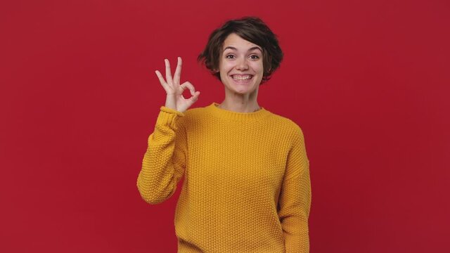 Smiling fun young woman 20s years old in yellow sweater posing isolated on red wall background in studio. People lifestyle concept. Look camera approvingly say wow it is ok showing okay sign gesture