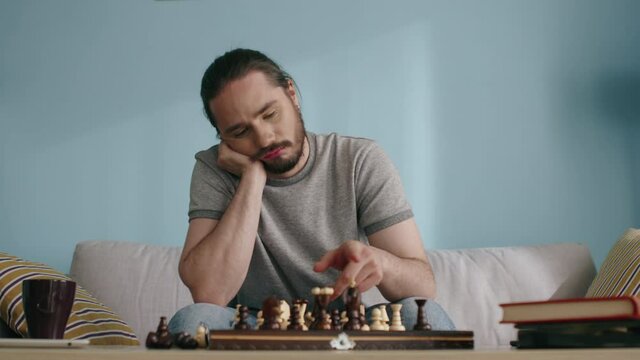 Young man is bored to play chess, looking sick and tired, killing time in self-isolation, missing live communication and his usual lifestyle, Slow motion.