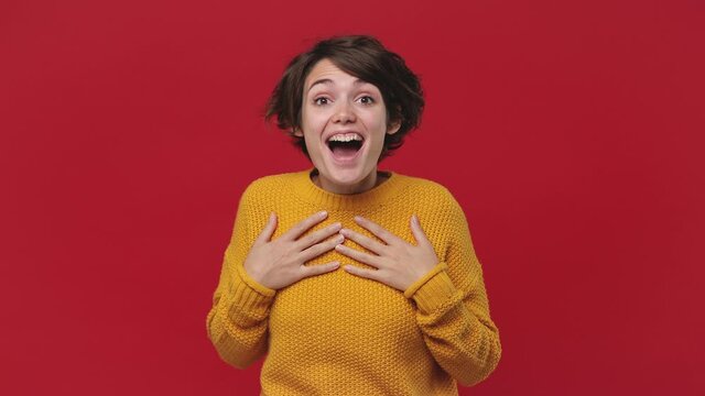 Smiling cute young woman 20s years old in yellow sweater isolated on red background studio. People lifestyle concept. Look camera ask who me oh it so sweet put hands on chest winner gesture say yes