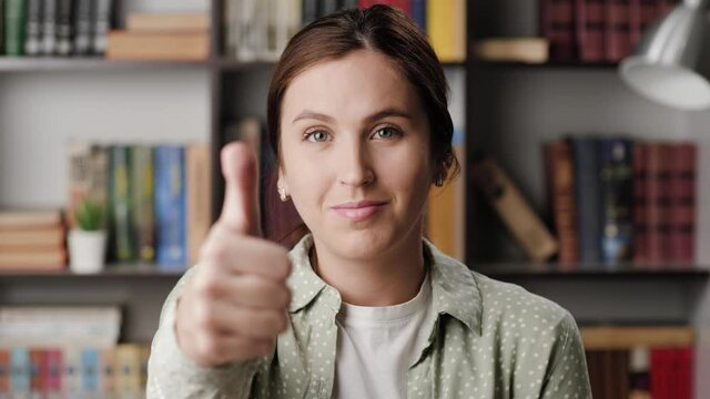 Woman thumb up. Positive smiling woman in office or apartment room looking at camera brings her hand up and shows her thumb up