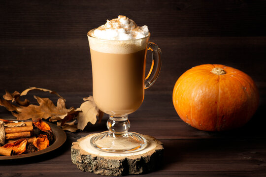 Pumpkin spice latte in a beautiful glass on a dark wooden background, next to it lies a pumpkin and a tray with spices.