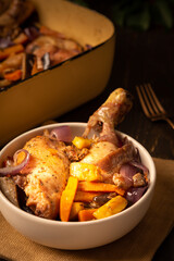 Plate with a serving of baked chicken with vegetables and iron brazier on a dark wooden background, chicken drumsticks with eggplant, pumpkin, onions and carrots baked in the oven