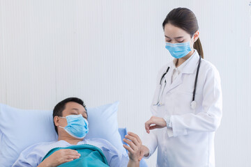 An Asian woman doctor is taking the pulse on the wrist of a man patient to check heart rate at the hospital. They wear a medical face mask to protect respiration system (Covid-19).  