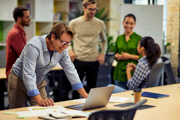 Happy businessman standing in the modern coworking space, looking at laptop screen and smiling while his young multiracial colleagues communicating on the background