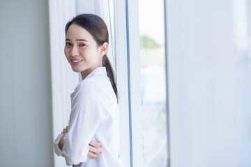 Asian beautiful woman in white shirt is standing lean against with window and white curtain.
