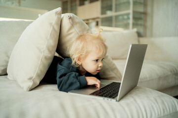 plays educational smart children's games cartoons in the app on the website on your computer laptop. little smiling blonde girl with a ponytail. sitting on the couch watching the monitor