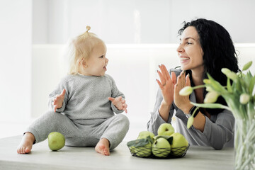 Fototapeta premium daughter Babes baby and mom brunette young family play smiling happiness in the kitchen. Clothing comfortable monochrome stylish, green apples on the table, modern bright kitchen design