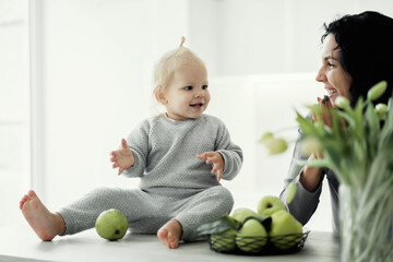 Fototapeta premium portrait of a young mother brunette and daughter blonde kids playing in the kitchen with green apples. On the table there are flowers, tulips, bright, stylish modern kitchen