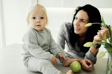Fototapeta premium little cute daughter girl kids baby blonde and mom brunette, young family playing smiling happiness in the kitchen, holding a green Apple playing in their hands. modern light kitchen design