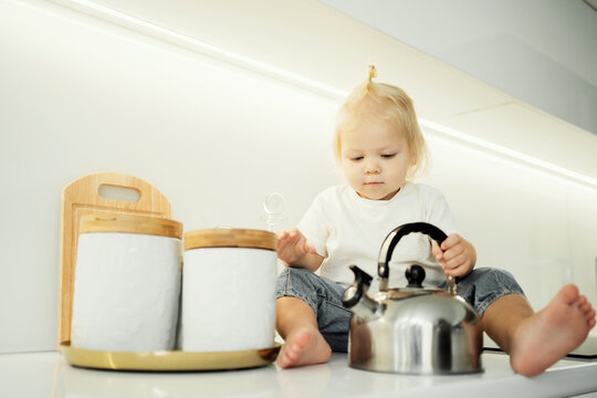 little girl baby kids sitting on the table and playing with dishes kettle metellic, stylish clothes, ponytail hair, jeans on her feet. photos for children's companies, products, and ads.