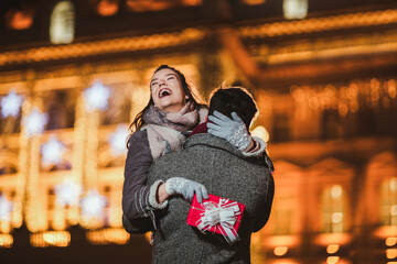 Young couple outdoor with holiday's brights in background. Man presenting gift to woman.