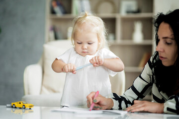 she was wearing a white shirt and blue jeans. white apartment design, daylight in the window. happy playful fun portrait girl blonde baby kids drawing on paper pictures