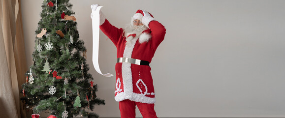 santa claus dressed in a red suit stands near the christmas tree. Watch the big list, put your hand on your head.