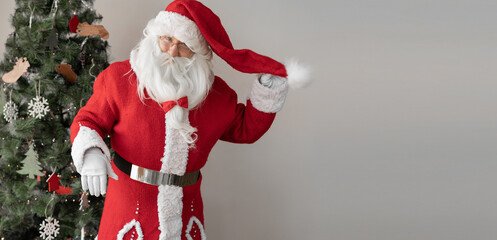 santa claus dressed in a red suit stands near the christmas tree. hand holds his cap.