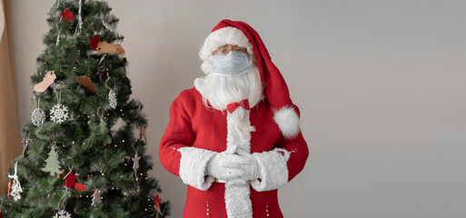 santa claus dressed in a red suit stands near the christmas tree. a protective medical mask is worn on the face.