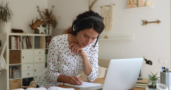35s woman lean at table wear headset looks at laptop screen writing down received information, listen e-teacher, gain new knowledge by videocall. Take part in webinar conducted over internet concept