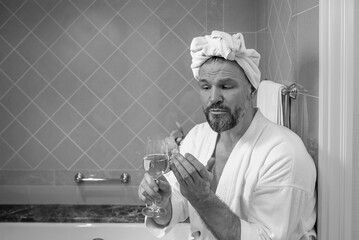 handsome mature man (gay) in white bathrobe and towel on his head sitting on bathroom with foam and...