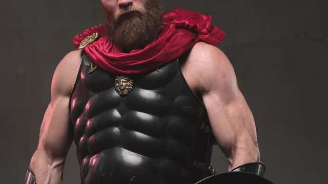 Camera's shooting brutal and muscular gladiator with dark armour and red mantle he stared upon to camera with ungentle looks down from above in dark background in studio.