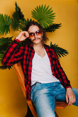 An attractive man with long hair and a mustache in a red plaid 80s disco shirt sits on a chair...