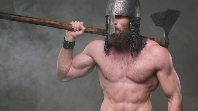 Muscular and shirtless warlike viking with helmet and axe on his shoulder poses rotating his head and looking around in smokey background then takes axe out of his shoulder holding it with his arms.