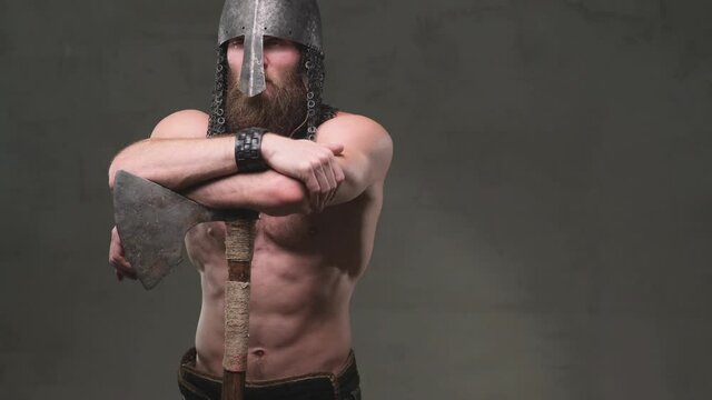Camera moves left from right shooting scandinavian barbarian with naked torso and muscular build he stays with crossed arms and poses with axe in dark background.