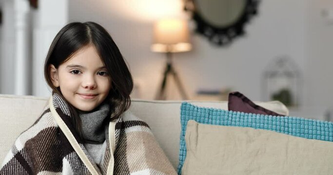Close up of pretty cute happy sick little female teenager smiling to camera while sitting at home alone. Small ill girl in warm plaid in cozy room. Kid with virus infection. Pandemic. Portrait concept