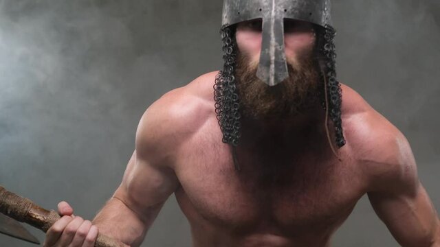 Naked and savage scandinavian barbarian with muscular build and helmet looks around holding his axe and searching for enemies in smokey background in studio.