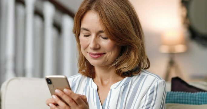 Close up portrait of pretty cheerful woman in good mood typing on smartphone indoors. Happy beautiful Caucasian middle-aged female smiling while texting on cellphone at home. Leisure concept