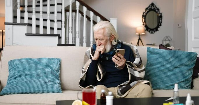 Caucasian old grey-haired sick man in plaid sitting on sofa in room and coughing while typing on smartphone. Unwell senior male havingvirus infection and browsing on cellphone at home. Health concept