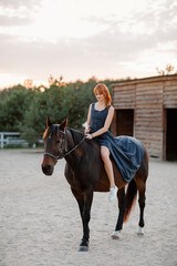 A beautiful red-haired girl in a dress walks with a horse at the horse ranch. Portrait of a girl with a horse. Girl riding a horse
