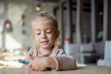 Little adorable blonde girl in cafe playing games on smartphone