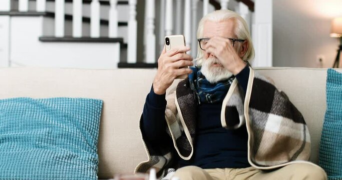 Portrait of sick grey-haired man sitting in cozy room and videochatting on cellphone. Senior male pensioner in warm plaid talking on video on smartphone at home indoors. Virus infection Health concept