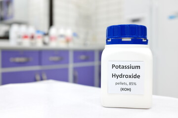 Selective focus of a bottle of pure potassium hydroxide or KOH chemical compound beside a petri dish with white solid pellets. Chemistry research laboratory background with copy space.