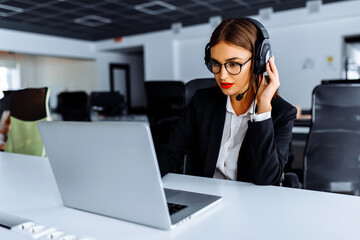 Smiling young female operator in headphones with headset working with laptop sitting at table at workplace, technical support concept