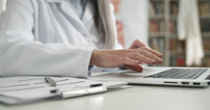 Crop view of woman in white professional coat typing and filling in medical documentation. Female doctor working with laptop in office while sitting at table. Concept of routine and work
