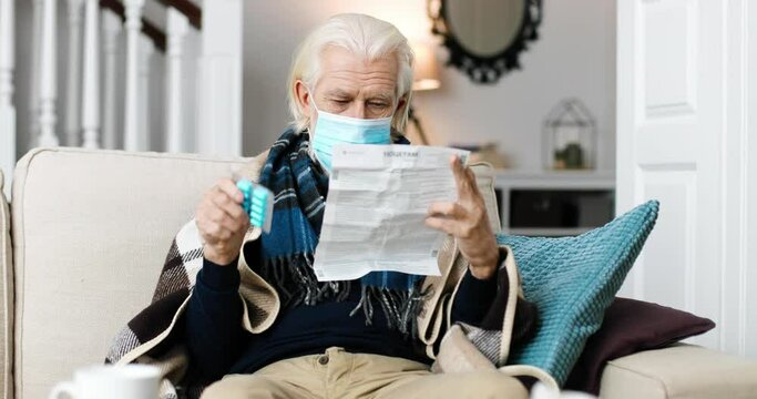 Portrait of senior Caucasian male in mask sitting at home and reading prospectus for pills. Sick unwell old male holding medicine in hand in room. Pandemic virus concept. Disease treatment